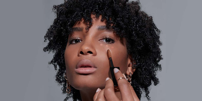 Woman with dark skin tone showing how to apply all over concealer under the eyes.